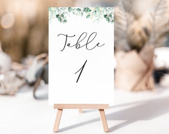 Peut inclure: Une carte blanche avec un feuillage vert en haut, indiquant "Table 1" en calligraphie noire. La carte est affichée sur un petit chevalet en bois.