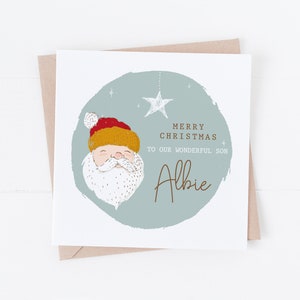 Puede incluir: Una tarjeta de Navidad con una ilustración de dibujos animados de Papá Noel. La tarjeta tiene un fondo azul con estrellas blancas y el texto "Feliz Navidad a nuestro maravilloso hijo Albie".