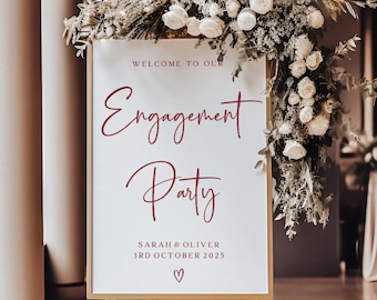 May include: A white engagement party sign with burgundy lettering, framed in gold, and adorned with white flowers and greenery. The sign reads "Welcome to Our Engagement Party" with the names Sarah & Oliver and the date 3rd October 2025.