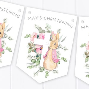 Puede incluir: Una pancarta blanca con un diseño floral rosa y una cruz blanca. Un conejo marrón con una chaqueta rosa está de pie frente a la cruz. La pancarta dice "Bautizo de May".