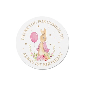Op de afbeelding: Een ronde witte sticker met gouden tekst en een aquarel illustratie van een konijn met een roze ballon. De tekst luidt "THANK YOU FOR COMING TO ALBA'S 1ST BIRTHDAY". Het konijn draagt een roze jas.