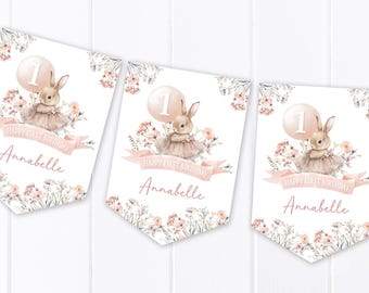 Op de afbeelding: Drie wimpelbanners met een aquarelontwerp voor een eerste verjaardag. Elke banner toont een konijntje met een tutu, een ballon met het nummer 1 en de naam "Annabelle". De banners zijn versierd met bloemelementen en de tekst "HAPPY FIRST BIRTHAY".