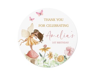 May include: A round sticker featuring a watercolor illustration of a fairy with brown hair and a yellow dress standing on a mushroom. The text reads THANK YOU FOR CELEBRATING Amelia's 1ST BIRTHDAY, accompanied by pink butterflies, yellow flowers, and a snail.