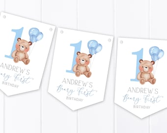 May include: Three white pennant banners with blue and white balloons, a brown teddy bear, and the text "Andrew's Beary First Birthday".
