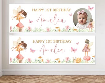 May include: A horizontal banner featuring the text HAPPY 1ST BIRTHDAY Amelia in gold and pink script. The design includes watercolor illustrations of a fairy on a mushroom, pink butterflies, green foliage, and a circular photo of a baby.
