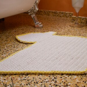 May include: A white bath mat with a yellow border in the shape of a T-shirt. The mat is on a brown and white tiled floor.