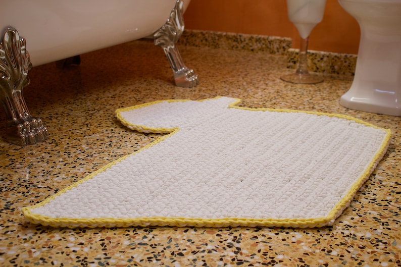 May include: A white bath mat with a yellow border in the shape of a T-shirt. The mat is on a brown and white tiled floor.