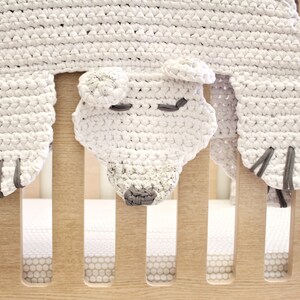 May include: White crocheted bear head crib toy with grey accents. The bear is hanging from a wooden crib rail.