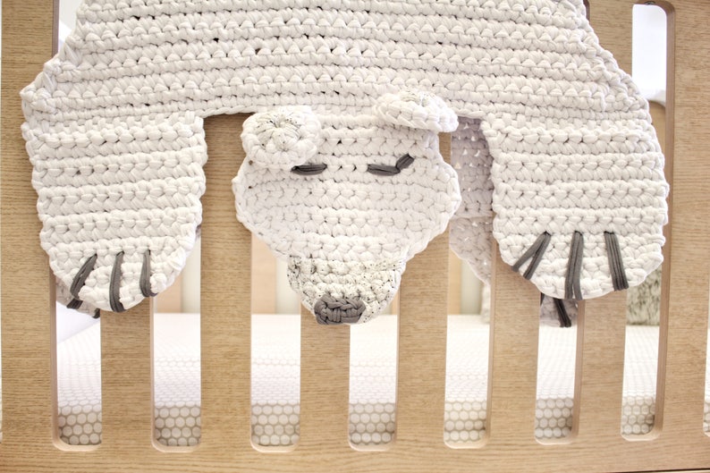 May include: White crocheted bear head crib toy with grey accents. The bear is hanging from a wooden crib rail.