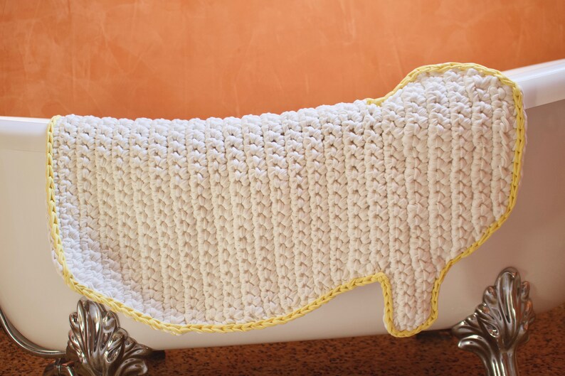 May include: White crocheted bath mat with a yellow border. The mat is shaped like a foot and is placed in a white bathtub.
