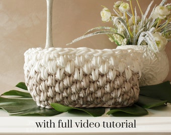 Op de afbeelding: Een gehaakte mand met een witte bovenkant en een beige onderkant. De mand staat op een groen blad. De tekst "with full video tutorial" staat onder de mand.