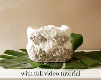 May include: A handmade, square crochet basket in white and tan yarn. The basket is sitting on a large green leaf. The text "with full video tutorial" is at the bottom of the image.