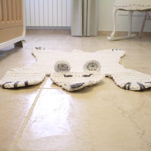 May include: A white crocheted polar bear rug with grey felt accents for the eyes and nose. The rug is laid out on a light beige tiled floor.