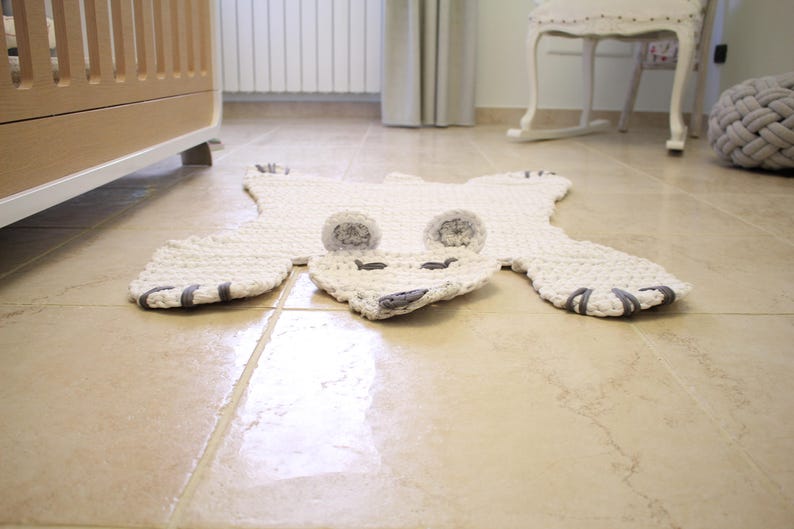 May include: A white crocheted polar bear rug with grey felt accents for the eyes and nose. The rug is laid out on a light beige tiled floor.
