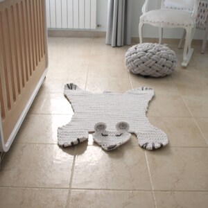 May include: A white crocheted rug shaped like a bear with black accents. The rug is on a beige tiled floor.