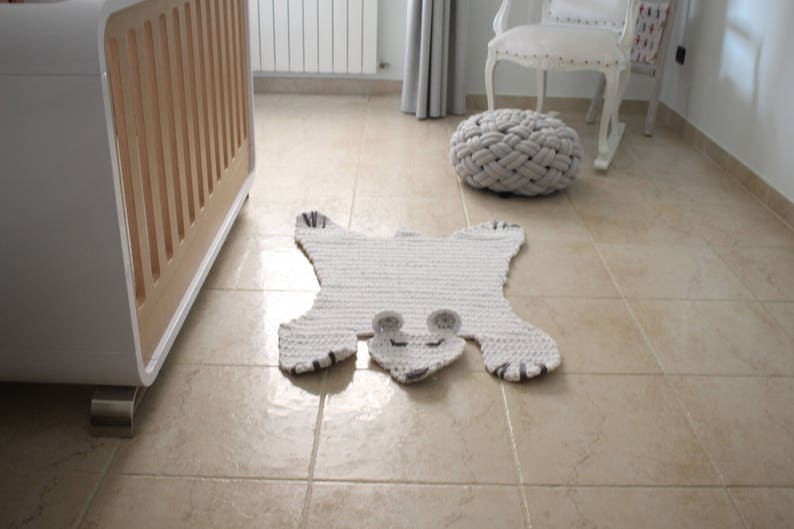 May include: A white crocheted rug shaped like a bear with black accents. The rug is on a beige tiled floor.
