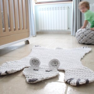 May include: A white crocheted rug shaped like a bear with grey accents. The rug is on a light brown tiled floor.