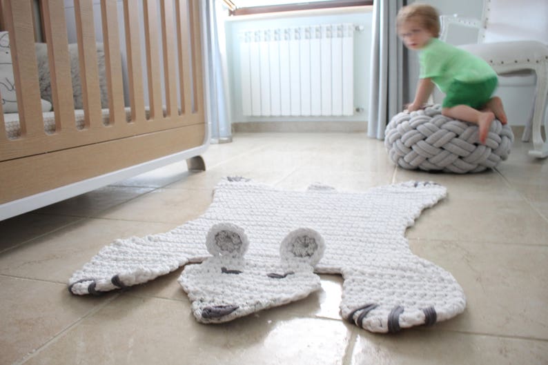 May include: A white crocheted rug shaped like a bear with grey accents. The rug is on a light brown tiled floor.