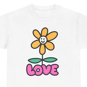 Könnte beinhalten: Weißes T-Shirt mit einer gelben Blume mit einem Smiley-Gesicht und grünen Blättern. Das Wort "LOVE" ist in rosa Blasenbuchstaben unter der Blume geschrieben.