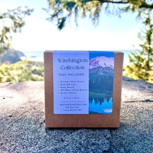May include: A brown box with a label that says "Washington Collection" and lists the teas included: Olympic Mint Green, Emerald City, Ruby Beach, Whidbey Island Breeze, and Ocean Shares. The label also includes the weight of the tea, 12 oz or 40 g, and the website address, BEACHHOUSETEAS.COM. The box is sitting on a rock with a view of the ocean and mountains in the background.