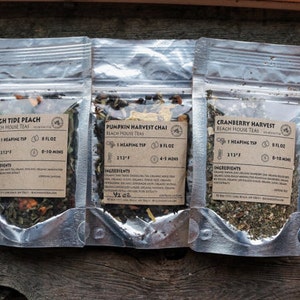 May include: Three small, clear plastic pouches containing loose leaf tea blends. The pouches are labeled with the names "High Tide Peach", "Pumpkin Harvest Chai", and "Cranberry Harvest". Each label includes the brand name "Beach House Teas", the ingredients, brewing instructions, and serving size.