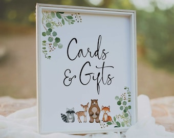 May include: White sign with a wood frame and green foliage border. The sign says "Cards & Gifts" in black cursive font. There are four woodland animals at the bottom of the sign: a raccoon, a deer, a bear, and a fox.