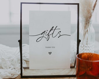 May include: A black framed sign with the word "gifts" written in cursive script. The sign also includes the text "Thank You" and a small heart symbol. The sign is sitting on a wooden table with a glass vase and a candle holder.