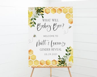 May include: A gender reveal sign on an easel with the text "What Will Baby Bee?" and "Welcome to Matt & Lauren's Gender Reveal 05.29.2021". The sign features a honeycomb design, watercolor bees, and floral accents.