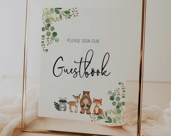 May include: A gold framed sign with a white background and green foliage border. The sign says "Please sign our Guestbook" with a cute illustration of woodland animals including a raccoon, deer, bear, and fox.
