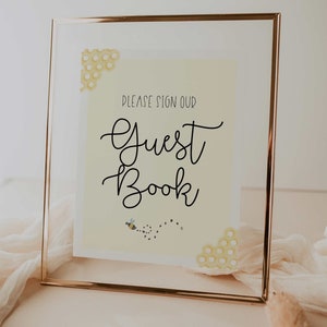 Puede incluir: Un cartel con marco dorado y fondo amarillo con el texto "Por favor, firme nuestro libro de visitas" en letra cursiva negra. El cartel presenta una peque&ntilde;a ilustraci&oacute;n de abeja y patrones de panal.