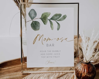 Pode incluir: Uma placa dourada enquadrada com uma ilustração em aquarela de um ramo de eucalipto. A placa diz "Mom-osa Bar" e inclui instruções para fazer uma mimosa: "Despeje o espumante, adicione um pouco de suco, cubra com fruta. Saúde!"