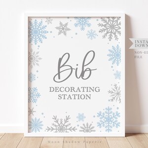May include: A printable sign with a white background and a gray border. The sign has a blue and silver snowflake design around the border. The text on the sign reads "Bib Decorating Station".