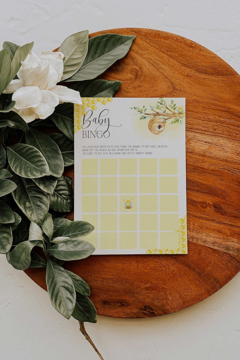 May include: A yellow and white baby shower bingo card with a bee illustration. The card has the text "Baby Bingo" and instructions to fill each box with gifts you think the mama-to-bee will receive. Mark off the boxes as she opens her gifts. The first to get five in a row and yells "Bingo" wins.