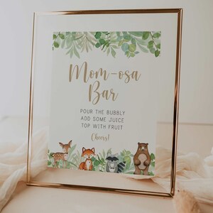 May include: A framed sign with a green leafy border and the text "Mom-osa Bar" in gold lettering. The sign instructs to pour bubbly, add juice, and top with fruit.  The bottom of the sign features a cartoon illustration of a deer, fox, raccoon, and bear.
