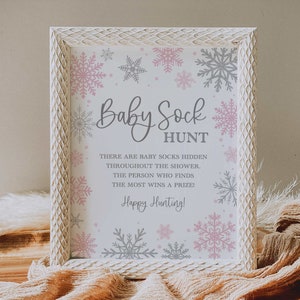 May include: A white frame with a pink and gray snowflake design. The text inside the frame reads "Baby Sock Hunt" and "There are baby socks hidden throughout the shower. The person who finds the most wins a prize! Happy Hunting!"