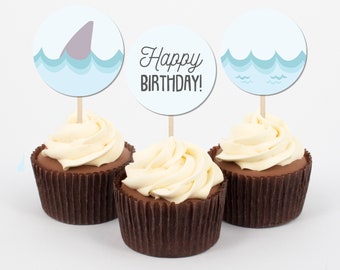 May include: Three cupcakes with blue and white frosting and toppers. The toppers are round and feature a shark fin, waves, and the text "Happy Birthday!"
