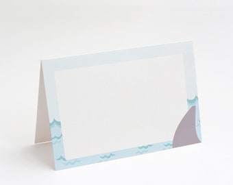 May include: A light blue place card with a white border and a shark fin design. The border is decorated with a repeating pattern of blue waves.