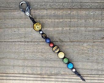 May include: A beaded keychain with a silver clasp. The keychain features a series of colorful, round beads in various colors, including yellow, blue, red, and green, strung on a black cord. The beads have different patterns and textures.