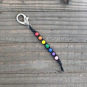 May include: A black macrame keychain with a silver clasp and a rainbow bead design. The beads are red, orange, yellow, green, blue, purple, and pink. The keychain is on a weathered wooden surface.