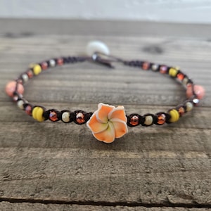 May include: A dark brown cord bracelet featuring a central peach-colored plumeria flower charm. The band is accented with a repeating pattern of small yellow, cream, and metallic orange beads, finished with a white button closure at the back.