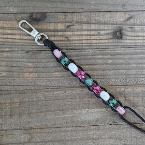 May include: Black lanyard with a silver clasp and a beaded section. The beaded section features pink, teal, and white beads. The lanyard is made of black cord and is designed to hold keys or an ID badge.