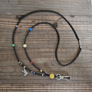May include: Black beaded lanyard with colorful accent beads. The lanyard features a silver astronaut charm and a silver clasp. The design includes yellow, blue, red, and green beads, creating a unique and eye-catching accessory.
