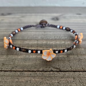 May include: A dark brown cord bracelet featuring three orange and white plumeria flower charms. The bracelet is accented with small white, orange, and dark brown beads arranged in a repeating pattern along the length of the cord.