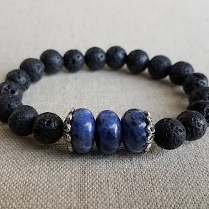 May include: A beaded bracelet featuring black lava rock beads and three blue sodalite beads. The blue beads are separated by silver-colored decorative beads. The bracelet is displayed on a neutral-colored surface.