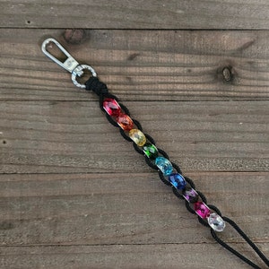 May include: A black macrame keychain with a silver clasp and a row of colorful, faceted beads in a rainbow pattern. The beads are red, orange, yellow, green, blue, indigo, and violet. The keychain is on a wooden surface.