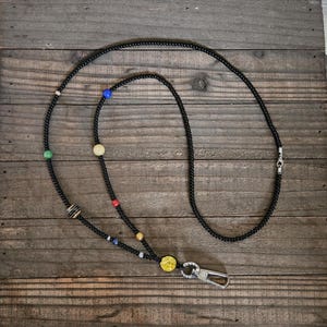 May include: A lanyard made of small black beads featuring a series of larger, multi-colored spherical beads arranged along the strand. The lanyard includes a silver-toned metal lobster clasp at the bottom for attaching items securely.