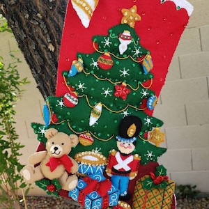 May include: A red felt Christmas stocking featuring a green felt Christmas tree, a teddy bear, a toy soldier, and presents. The stocking is adorned with sequins and beads.