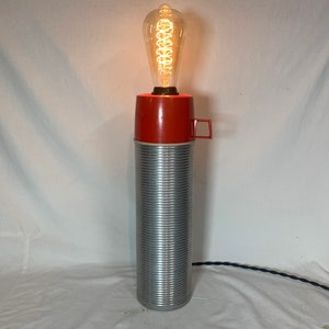 Könnte beinhalten: Eine Vintage-Lampe aus einem wiederverwendeten Thermos. Die Lampe hat ein rotes Oberteil und einen silbernen Korpus mit einer gerippten Textur. Sie verfügt über eine klare Glühbirne mit einem Filament.