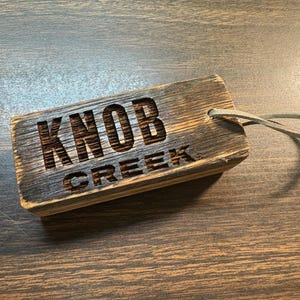Könnte beinhalten: Ein rechteckiges Holzschild mit dem eingravierten Text "KNOB CREEK". Das Schild besteht aus verwittertem Holz mit einer Lederschlaufe. Das Holz hat eine Mischung aus Braun- und Grautönen.