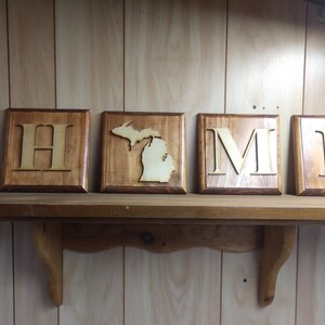 May include: Wooden sign spelling "HOME" with the letter "O" replaced by the outline of the state of Michigan. The sign is made of stained wood and has a rustic look.
